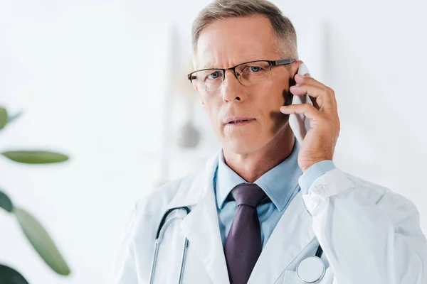 Красивий лікар дивиться на камеру і розмовляє по смартфону в клініці — стокове фото