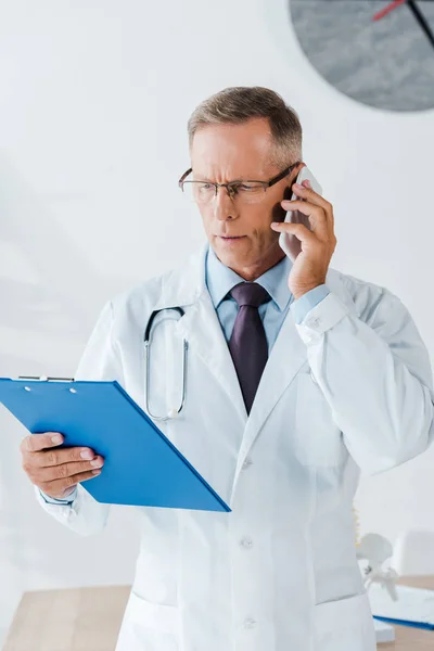 Серьезный врач в очках, глядя в буфер обмена и разговаривая на смартфоне — стоковое фото