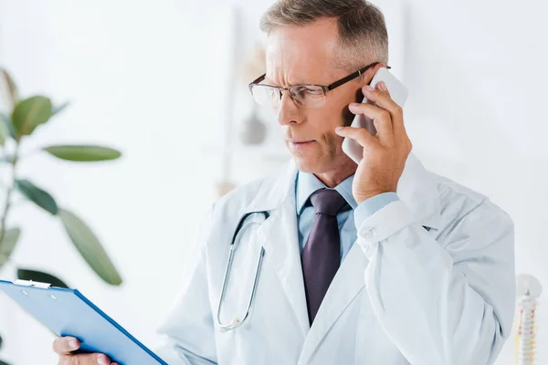 Serious doctor in glasses looking at clipboard while talking on smartphone — Stock Photo