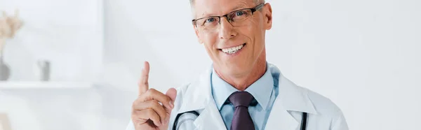 Panoramic shot of happy man in glasses pointing with finger in clinic — Stock Photo