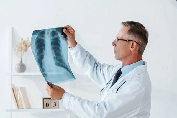Médico grave em casaco branco e óculos olhando para raio-x no hospital — Fotografia de Stock