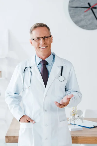Щасливий лікар в окулярах жестикулює, стоячи з рукою в кишені — стокове фото