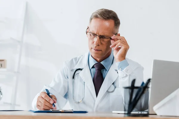 Selektiver Fokus eines gutaussehenden Arztes, der im Krankenhaus beim Schreiben auf Klemmbrett eine Brille berührt — Stockfoto