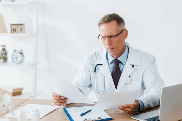 Guapo doctor en gafas mirando documentos en clínica - foto de stock