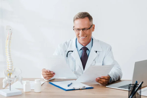 Zufriedener Arzt mit Brille beim Blick auf Dokumente in der Klinik — Stockfoto