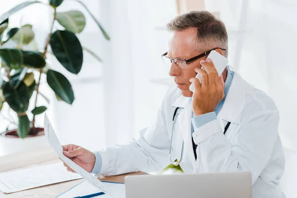 Arzt in Brille und weißem Kittel spricht auf Smartphone und schaut sich Dokument an — Stockfoto