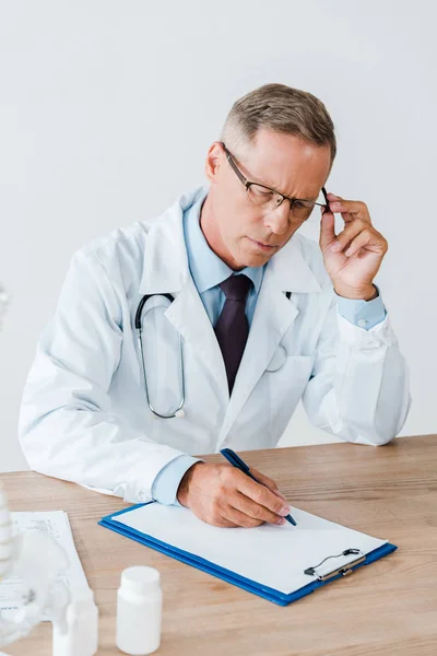Лікар в білому пальто торкається окулярів і пише діагноз в клініці — Stock Photo