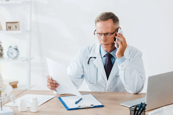 Docteur en lunettes et manteau blanc parlant sur smartphone et regardant du papier — Photo de stock