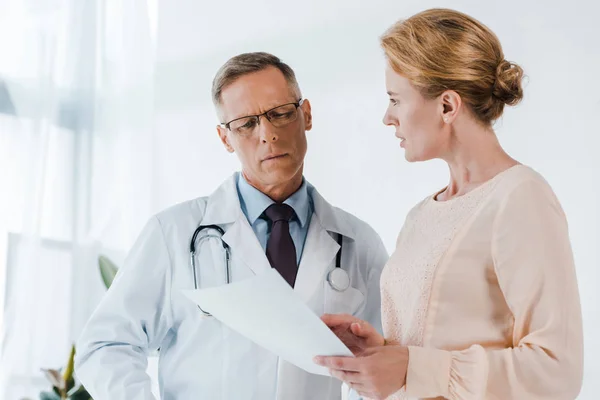 Médecin en lunettes et manteau blanc regardant document près de la femme — Photo de stock