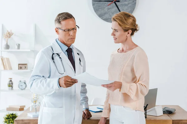 Arzt in Brille und weißem Mantel schaut sich Dokument neben attraktiver Frau an — Stockfoto