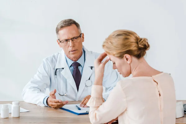 Selective focus of doctor in glasses looking at upset woman touching head while sitting near table — Stock Photo