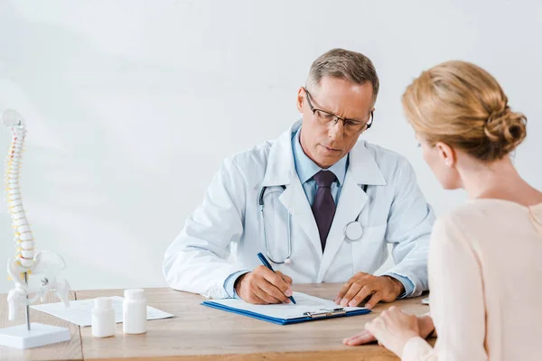 Selective focus of doctor in glasses writing diagnosis near woman — Stock Photo