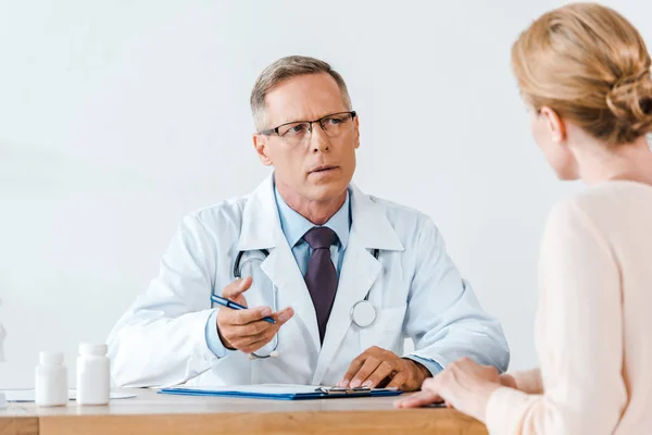 Selektiver Fokus des Arztes im weißen Kittel, der Frau ansieht und gestikuliert, während er am Tisch sitzt — Stockfoto