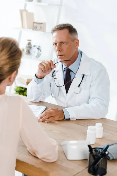Вибірковий фокус лікаря, який дивиться на жінку і тримає окуляри в клініці — стокове фото