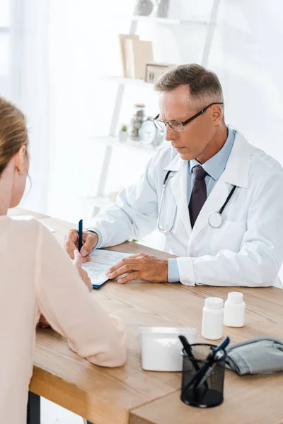 Ausgeschnittene Ansicht einer Frau in der Nähe eines Arztes mit Brille, die Diagnose schreibt — Stockfoto