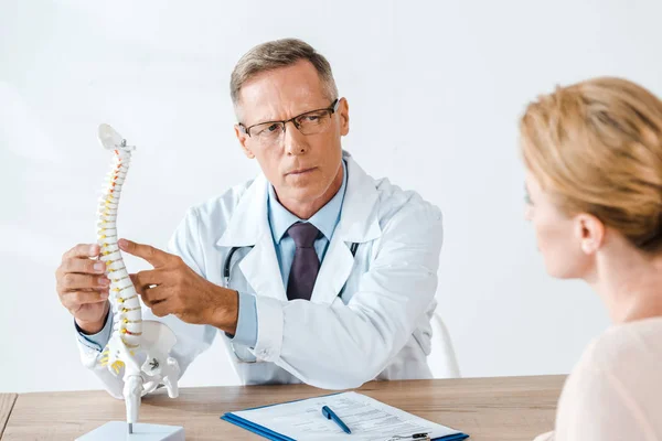 Selective focus of doctor in glasses pointing with finger at spine model near woman — Stock Photo