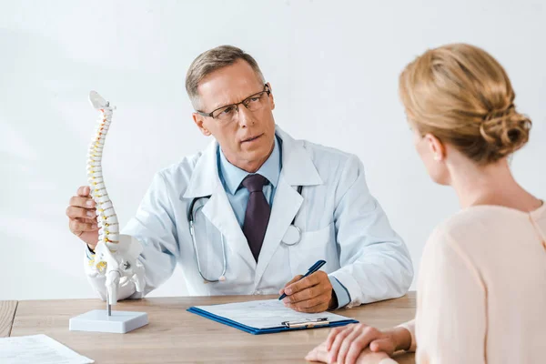 Selective focus of doctor in glasses touching spine model and looking at woman — Stock Photo