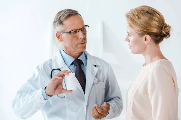 Médico en gafas gesto y sosteniendo botella cerca de la mujer en la clínica - foto de stock