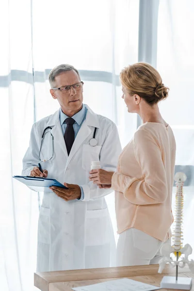 Doctor en gafas sujetando portapapeles con bolígrafo y mirando a mujer con botella - foto de stock