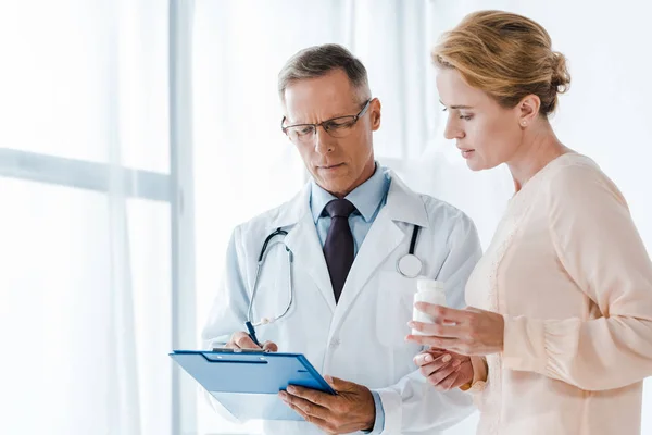 Doctor en gafas sujetando portapapeles con pluma cerca de mujer con botella - foto de stock