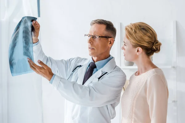 Красивый доктор в очках жестикулирует рядом с женщиной и смотрит на рентген — стоковое фото