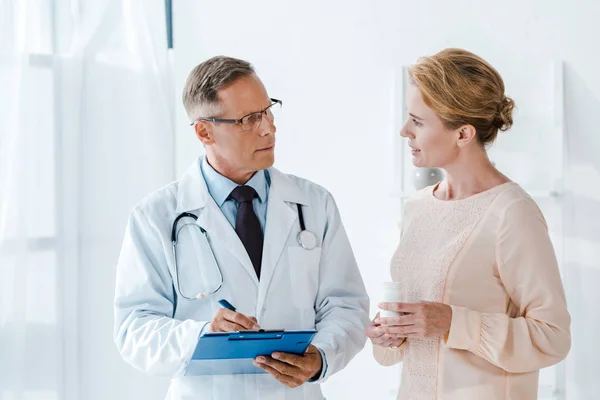 Doctor holding clipboard and pen while writing diagnosis and looking at woman with bottle — Stock Photo