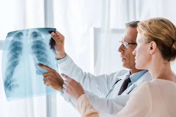 Schöner Arzt mit Brille zeigt mit dem Finger auf Röntgenbild in der Nähe attraktive Frau — Stockfoto