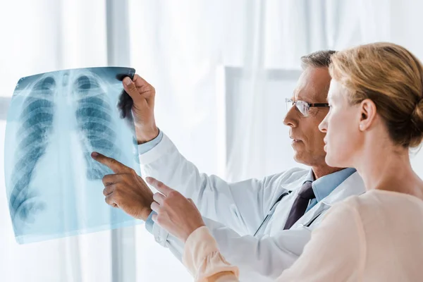 Guapo médico en gafas apuntando con el dedo a la radiografía cerca de atractiva mujer - foto de stock