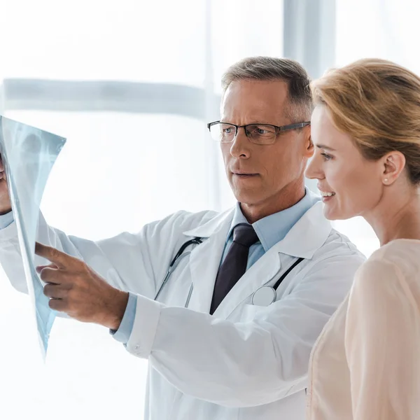 Gutaussehender Arzt mit Brille gestikuliert in der Nähe einer glücklichen Frau, während er Röntgenaufnahmen betrachtet — Stockfoto