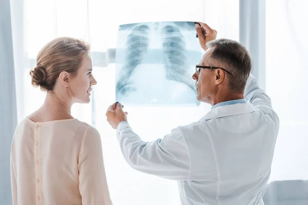 Médico em casaco branco e mulher olhando uns para os outros perto de raio-X na clínica — Fotografia de Stock