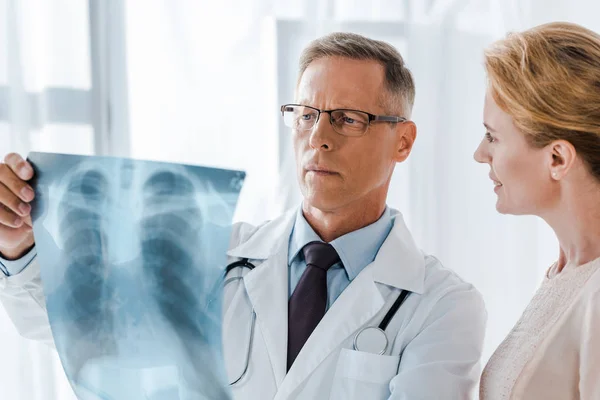 Mujer feliz mirando al doctor en bata blanca sosteniendo rayos X en la clínica - foto de stock