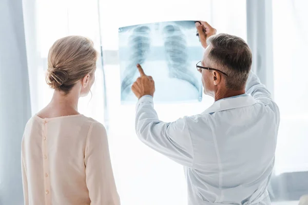 Médecin en manteau blanc pointant avec le doigt à la radiographie près de la femme à la clinique — Photo de stock