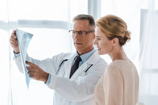 Arzt in weißem Mantel zeigt mit Finger auf Röntgenbild nahe positiver Frau in Klinik — Stockfoto