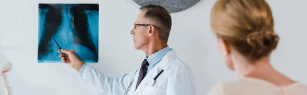 Panoramic shot of doctor in white coat holding pen near x-ray and woman in clinic — Stock Photo