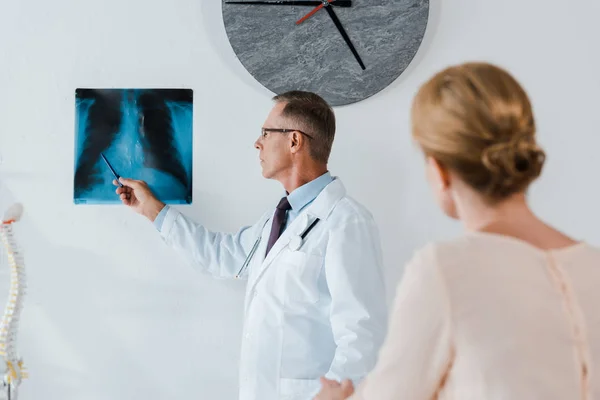 Enfoque selectivo del médico en la pluma de retención de capa blanca cerca de rayos X y la mujer en la clínica - foto de stock
