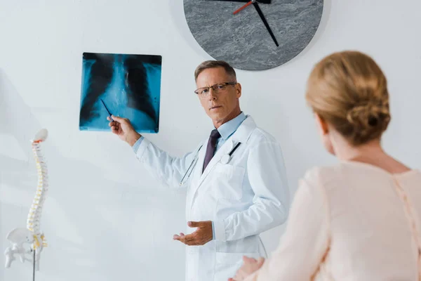 Foco seletivo do médico em casaco branco e óculos segurando caneta perto de raio-x e mulher na clínica — Fotografia de Stock