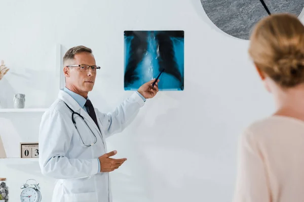 Selective focus of doctor in glasses holding pen near x-ray and woman in clinic — Stock Photo