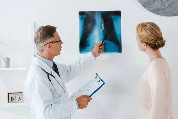 Лікар в окулярах тримає ручку біля рентгенівського знімка і пацієнта в клініці — стокове фото