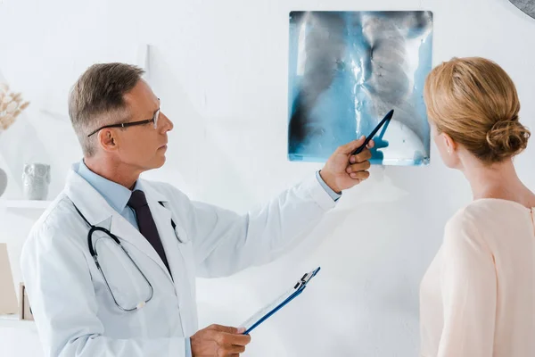 Doctor en gafas sosteniendo pluma cerca de rayos X y mujer en la clínica - foto de stock