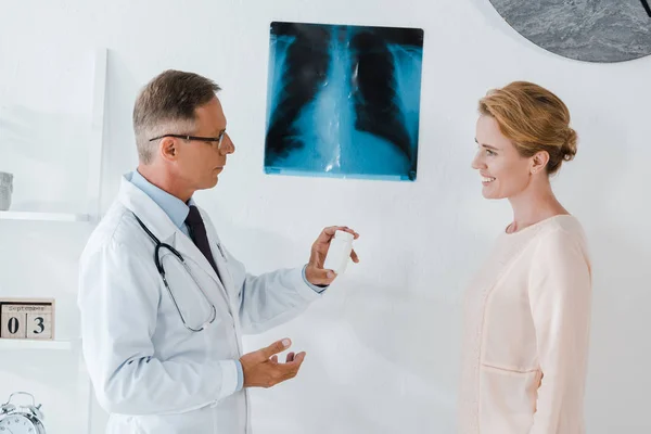 Beau médecin dans des lunettes debout près de rayons X et tenant bouteille près de femme gaie — Photo de stock