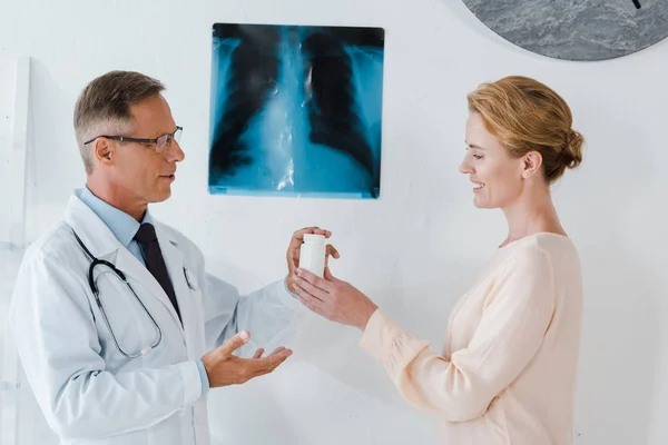 Handsome doctor in glasses standing near x-ray and giving bottle to cheerful woman — Stock Photo