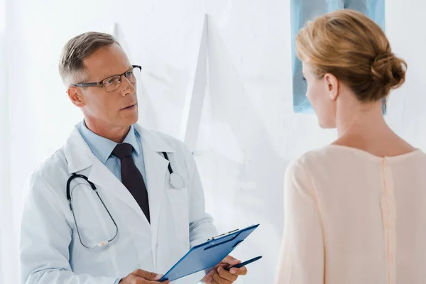 Doctor in glasses holding pen and clipboard near patient in clinic — Stock Photo