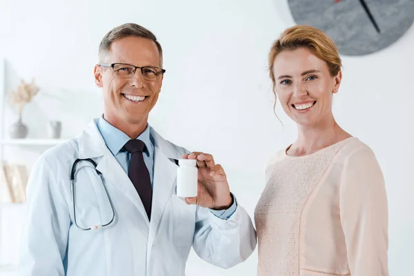 Fröhlicher Arzt in Brille lächelt, während er Flasche in der Nähe fröhlicher Patientin hält — Stockfoto