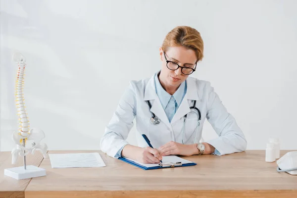 Attraente medico in occhiali scrittura diagnosi vicino modello di colonna vertebrale in clinica — Foto stock