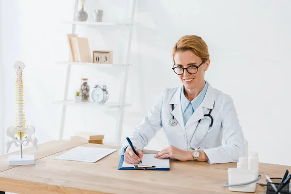 Happy doctor in glasses writing diagnosis near spine model in clinic — Stock Photo