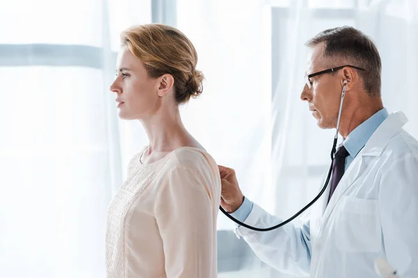 Vista laterale del medico con gli occhiali usando lo stetoscopio mentre esamina il paziente attraente in clinica — Foto stock