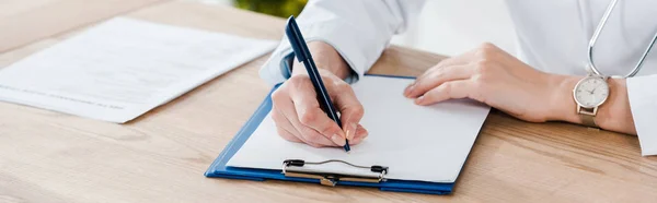 Panoramic shot of doctor writing diagnosis on wooden table — Stock Photo