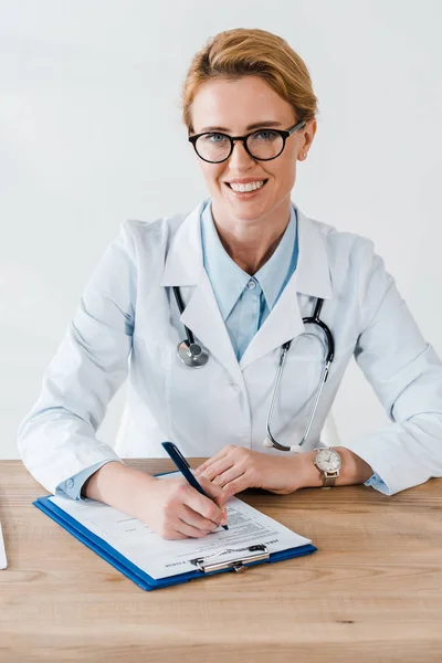 Glücklicher Arzt mit Brille lächelt, während er in die Kamera blickt und Stift in der Nähe des Klemmbretts hält — Stockfoto
