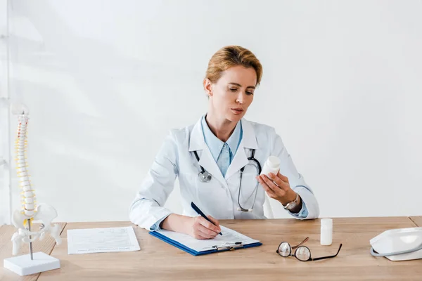 Attractive doctor holding bottle and pen near clipboard in clinic — Stock Photo