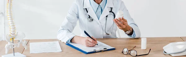 Panoramic shot of doctor holding bottle and pen near clipboard in clinic — Stock Photo